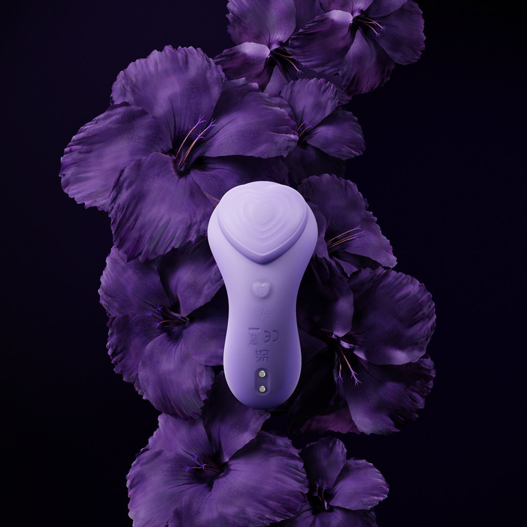 Hero shot of Velvet Rituals Whisper wearable vibrator in soft lavender, placed on a dark background with purple flowers, highlighting its elegant design and premium finish